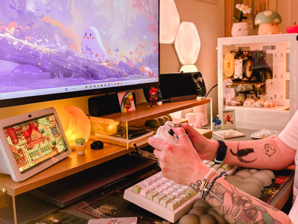 A gamer over 40 plays at their gaming desk with an X box controller. On the screen is a game with a ghost running up a tree trunk. The gaming desk is adorned with lights and trinkets. 