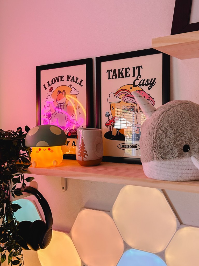 A shelf above the cosy gaming desk setup, holding two pictures, a light up mushroom toy, a narwhal plushie and headphones. 
