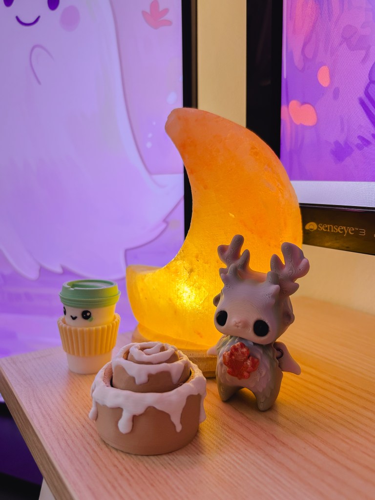 Small toys sitting on the cosy gaming desk infront of the monitor. There is a reindeer, a cinnamon bun, and a little toy cup of coffee.