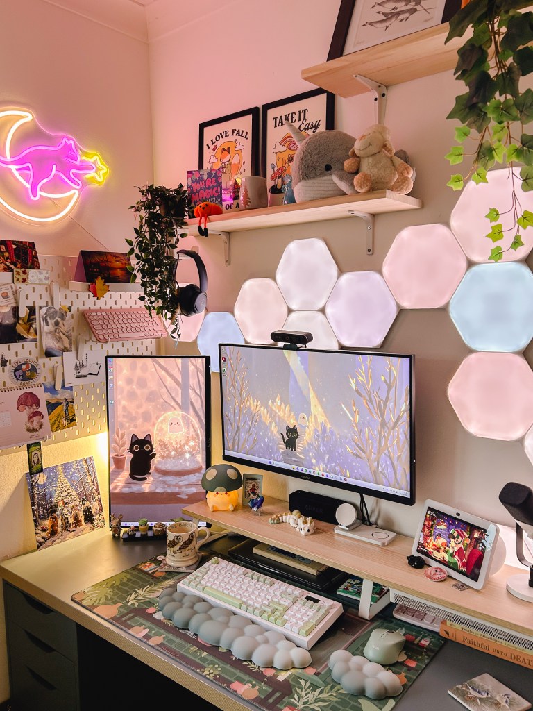 A cosy gaming desk setup for the winter. There are two monitors on the desk, pale blue and pink lights behind the screens, and two shelves holding plushes above the desk. 